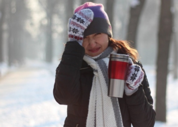 A women bundled up during a winter day with her hand on her head and holding a coffee.