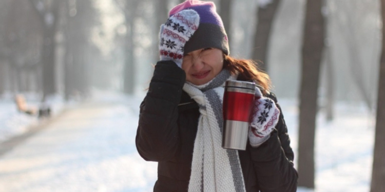 A women bundled up during a winter day with her hand on her head and holding a coffee.