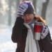 A women bundled up during a winter day with her hand on her head and holding a coffee.