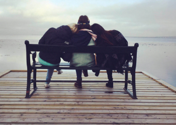 Three women sitting on a bench, leaning on each other while looking out at a lake.