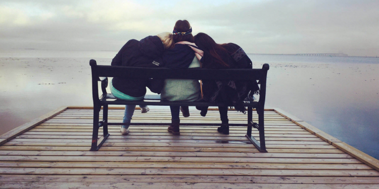 Three women sitting on a bench, leaning on each other while looking out at a lake.