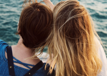 Two women sitting down and leaning on each other while they look out over the water.