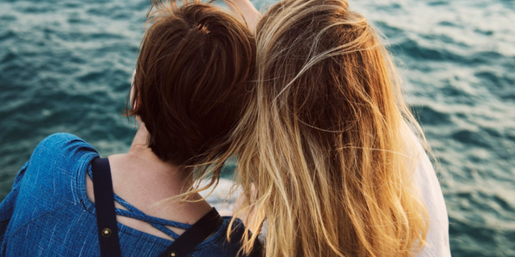 Two women sitting down and leaning on each other while they look out over the water.