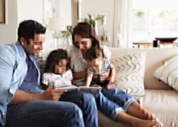 A mom and dad sit on a couch with their young girl and boy reading,