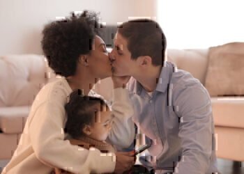 A mom holds her young daughter while holding her phone scrolling through social media and pauses to kiss her husband.