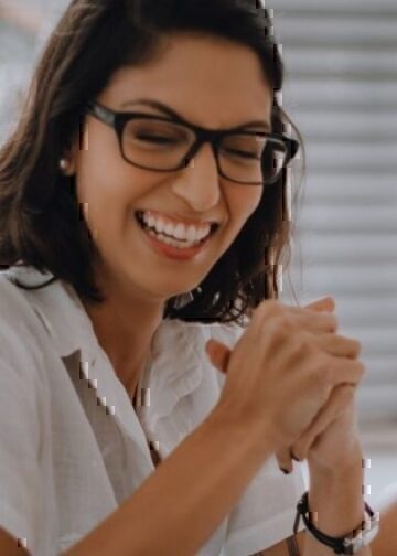 A women sitting down, while smiling and holding her hands together.