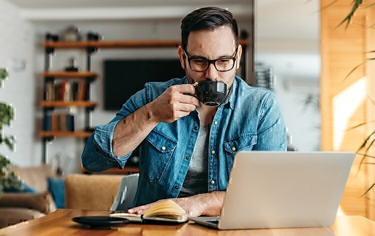 A man sipping coffee while completing school-work on his computer.