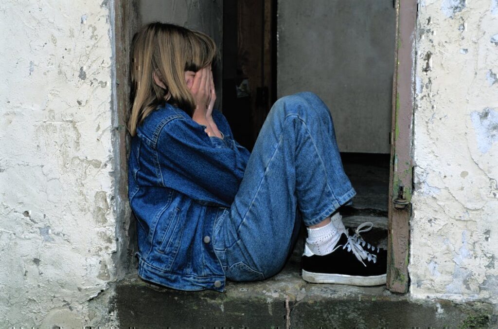 A young child sitting in a doorway while holding her face in her hands while crying.